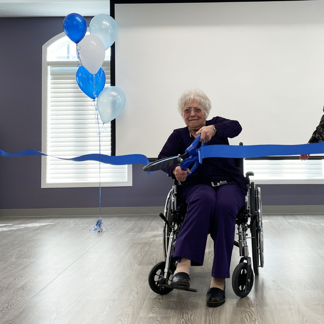 An elderly woman in a wheelchair cuts a ribbon with giant scissors 